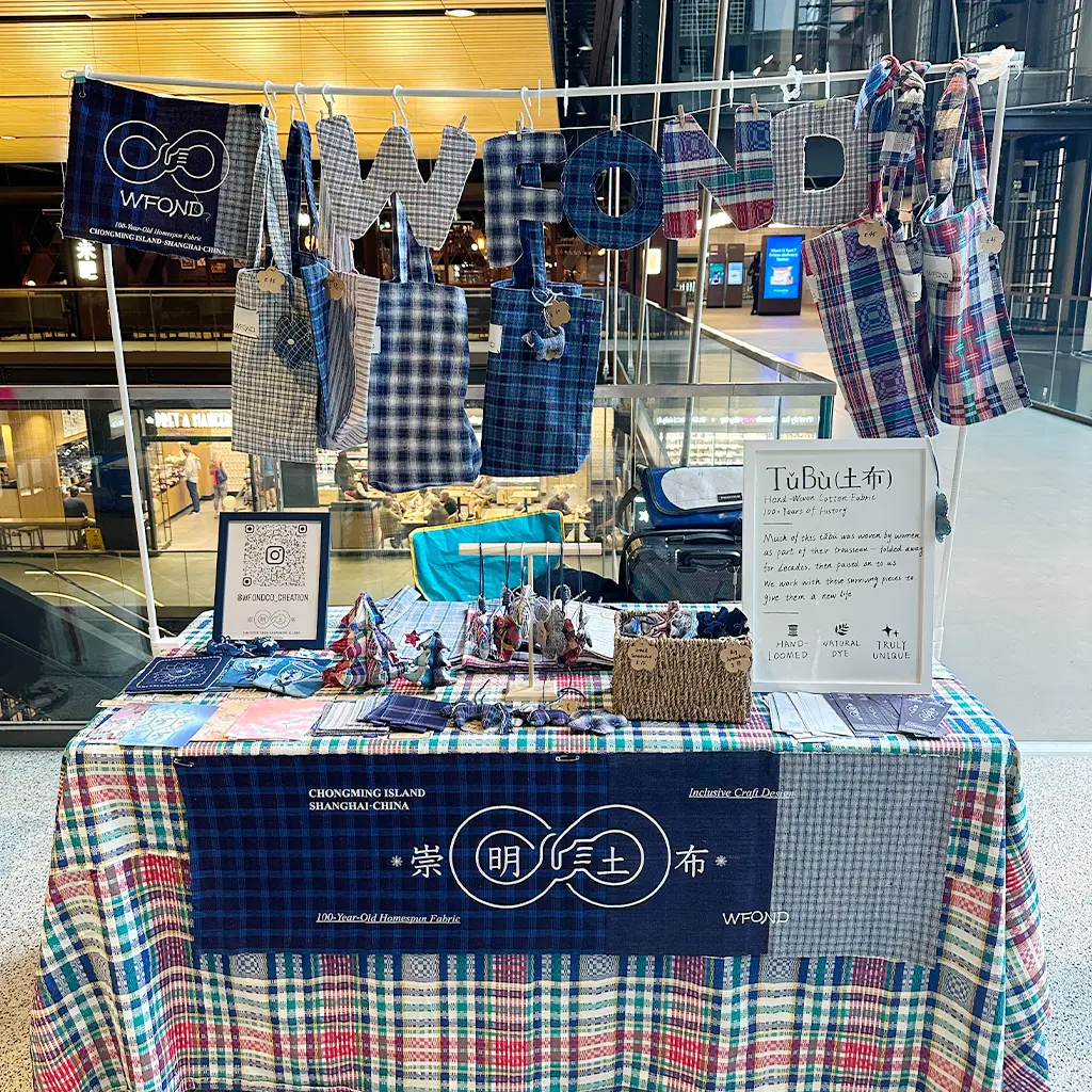 WFOND market stall at Battersea Power Station — Chongming Tubu totes and charms hanging from a line of hand-cut WFOND letters, above a checked cloth and a hand-lettered “Tǔbù — hand-woven cotton fabric” sign.