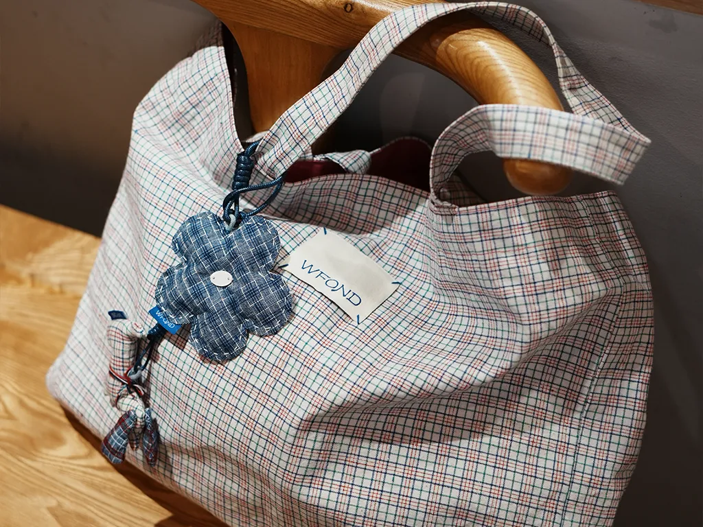 A checked Chongming Tubu tote bag with a denim-blue WFOND flower charm tied to the handle, hanging on a wooden stand.