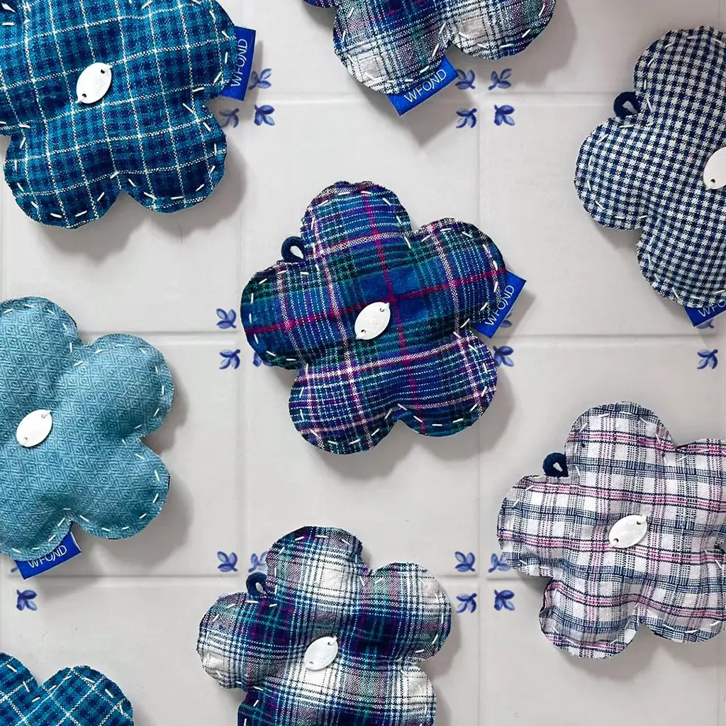 A grid of handmade WFOND flower charms in assorted blue plaid Chongming Tubu, hand-stitched with a shell-button centre, on a white tiled wall.