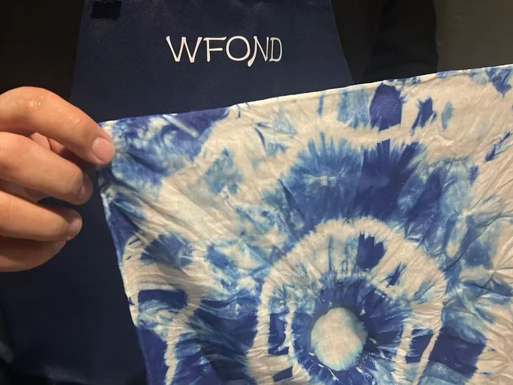 A maker holding up a finished indigo-dyed cloth with a circular sunburst pattern, a navy WFOND apron behind.